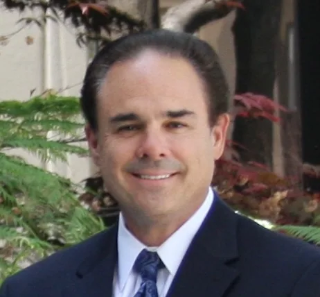 Man in dark blazer and blue tie smiling at camera with green foliage in background