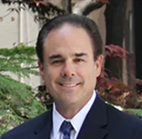 Professional headshot of a man in a dark blazer and tie smiling at the camera with green plants in the background